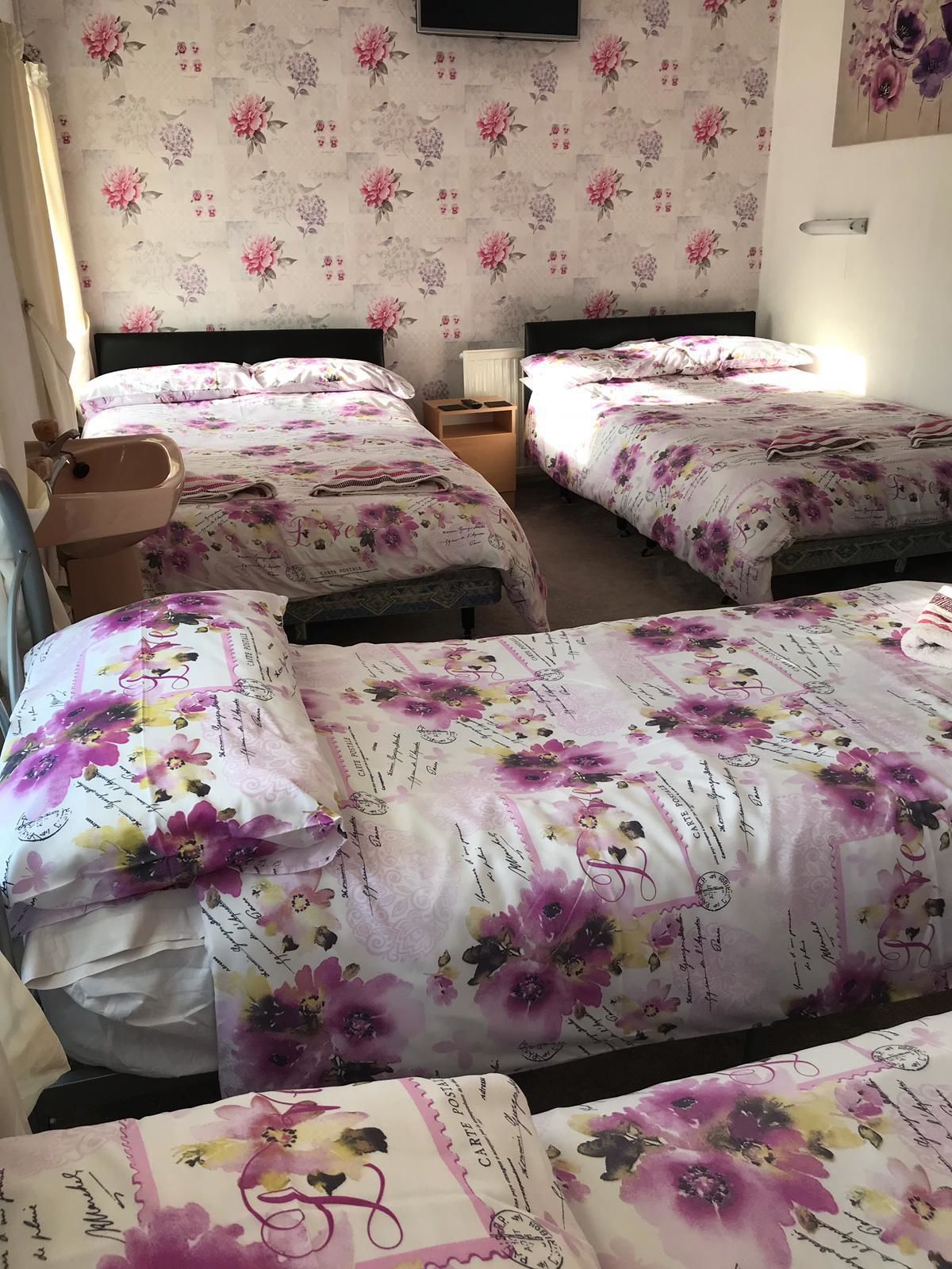 A hotel room with four beds, each with floral bedding. There is a television mounted on the wall and a small bedside table with a lamp.
