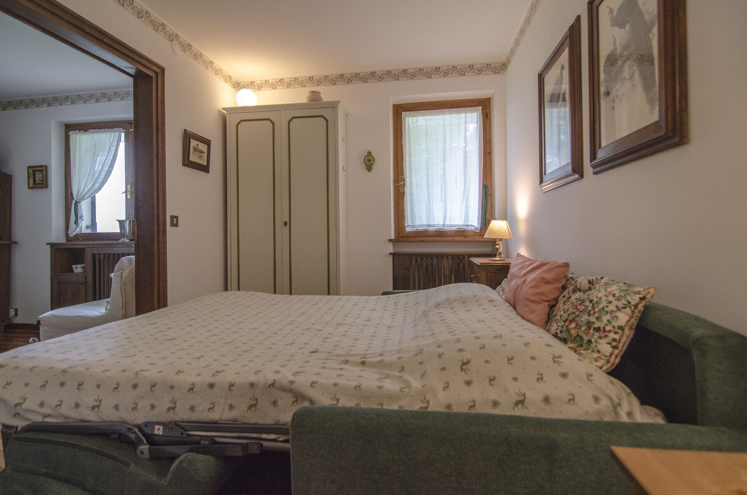A bedroom with a bed, a chair, a dresser, a window with curtains, and framed pictures on the wall.