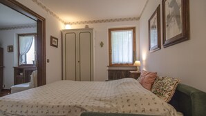 Une chambre à coucher comprenant un lit, une chaise, une commode, une fenêtre avec des rideaux et des tableaux encadrés accrochés au mur.