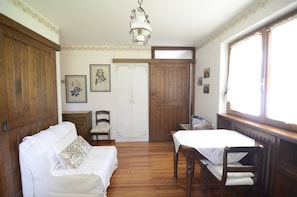 Une pièce avec un plancher en bois, un canapé blanc orné d’un coussin à motifs floraux, une table en bois recouverte d’une nappe blanche, une porte en bois et un luminaire suspendu.