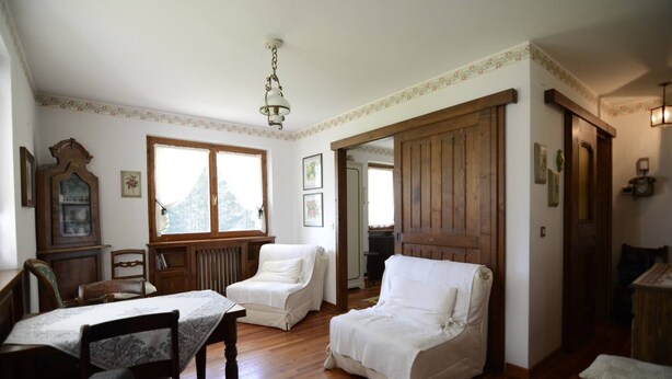 Une pièce avec un plancher en bois, une table à manger avec des chaises, un fauteuil blanc, un meuble en bois et une grande fenêtre.