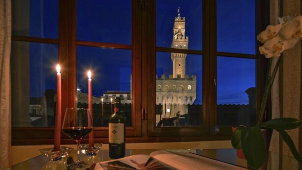 Une table avec des bougies, une bouteille de vin et un livre ouvert, près d’une fenêtre donnant sur une tour.