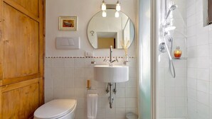 Une salle de bain avec des toilettes blanches, un lavabo avec un miroir au-dessus, une douche avec un pommeau de douche à main et une petite poubelle.