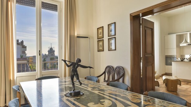 Ein Essbereich mit einem Marmortisch, Stühlen und einer Statue. An der Wand hängen gerahmte Bilder und durch das Fenster ist ein Blick auf Gebäude zu sehen.