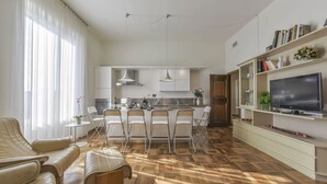 Eine moderne Küche mit Kochinsel, weißen Schränken und Pendelleuchten. Es gibt eine Essecke mit Stühlen und einer Couch.