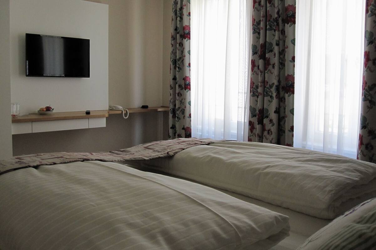 Ein Zimmer mit zwei Betten, einem an der Wand montierten Fernseher, einem Nachttisch mit Telefon und einem Fenster mit floralem Vorhang.