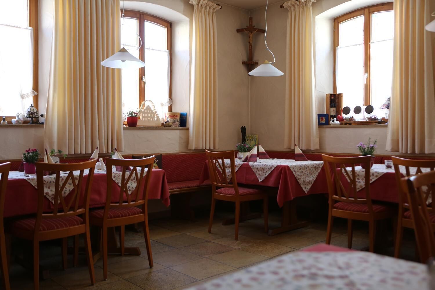 A dining area with wooden chairs and tables, large windows with curtains, and a cross on the wall.