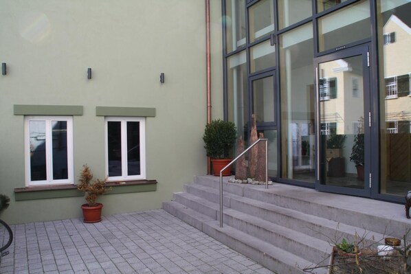 Entrada de un edificio con un conjunto de escalones de hormigón que conducen a una puerta de cristal, flanqueada por dos ventanas con marcos blancos y detalles verdes.
