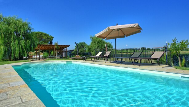 Ein klares, blaues Schwimmbecken mit Liegestühlen und Sonnenschirmen, umgeben von Bäumen und einer Pergola.