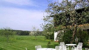 A garden with white metal chairs and tables, a tree with blossoms, and a building in the background. - Cafe Best Gästezimmer (Erbach)