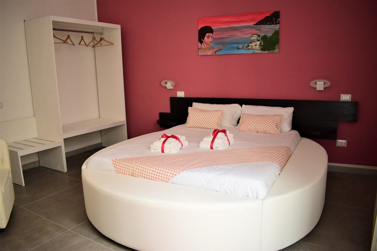 Una cama redonda con ropa de cama blanca y detalles en rojo, un armario blanco y un cuadro en la pared.