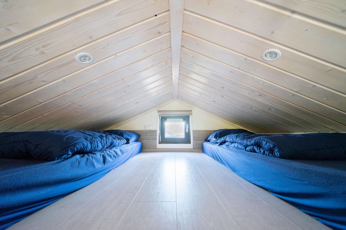 Une pièce avec un plafond en pente, deux lits avec une literie bleue, un puits de lumière et un plancher en bois.