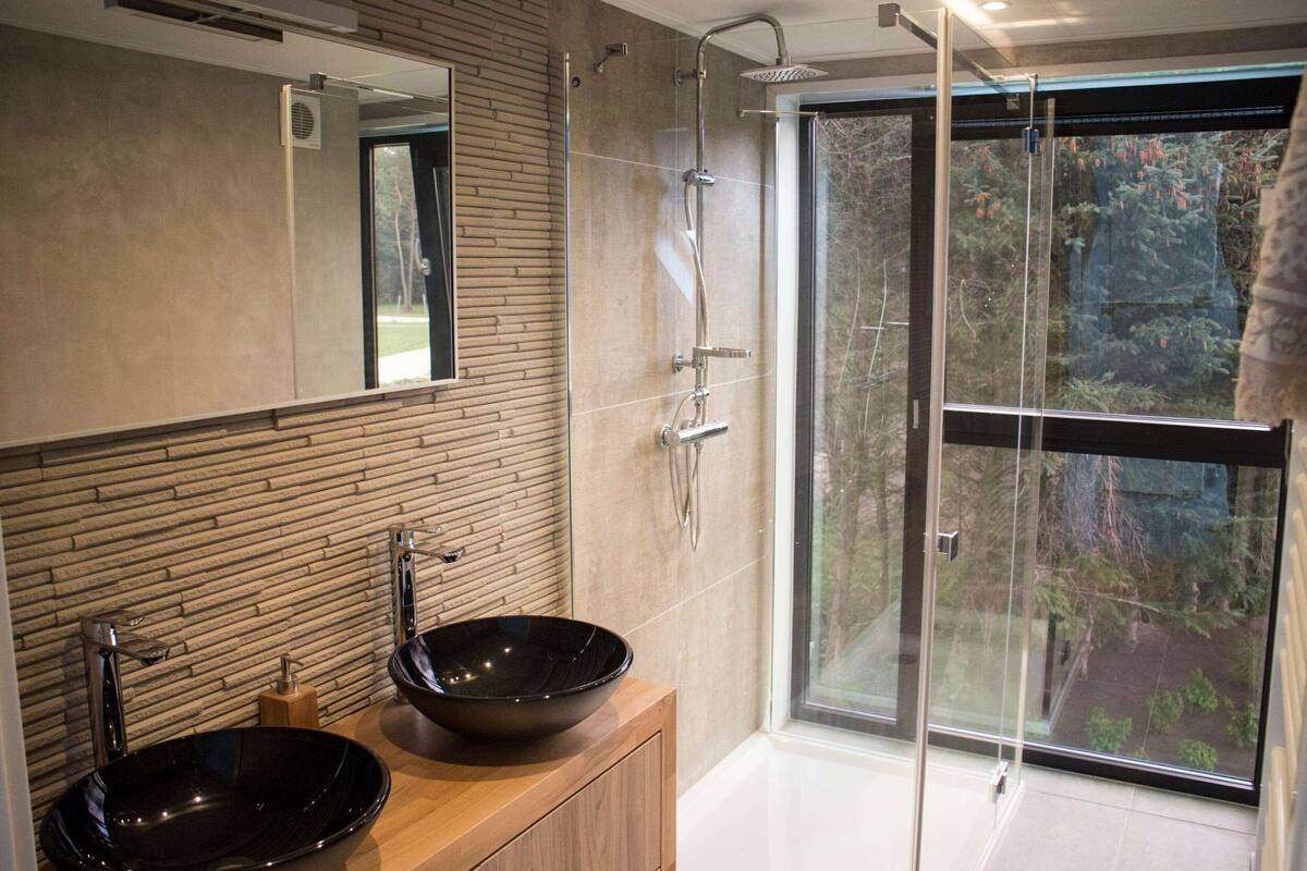 Une salle de bain moderne avec un grand miroir, deux lavabos, une douche et une porte en verre donnant sur une vue sur la forêt.