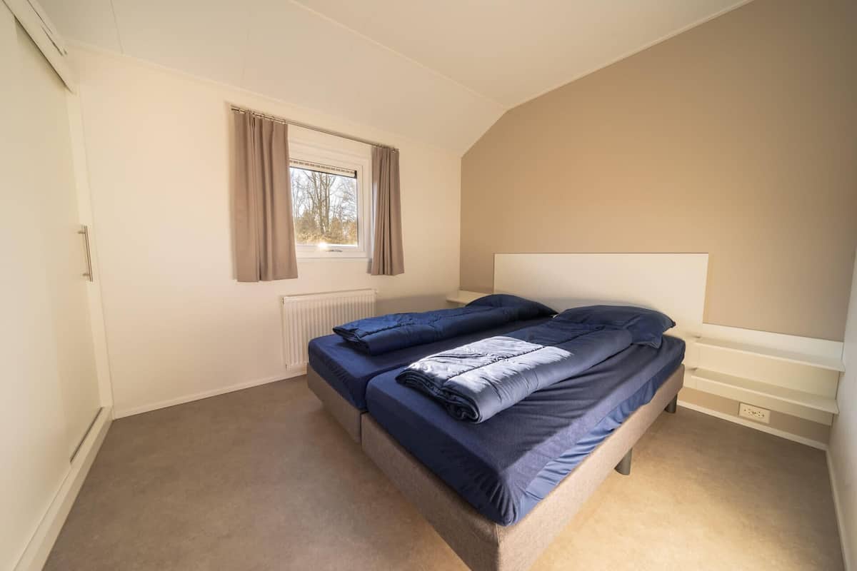 Une chambre à coucher avec un lit, une fenêtre avec des rideaux, un radiateur et une tête de lit blanche.