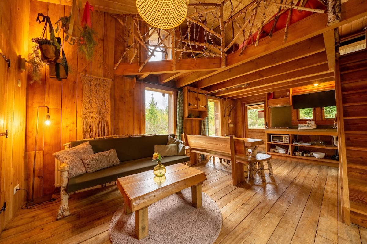 L’intérieur d’un chalet en bois comprend un canapé, une table en bois, un banc, un coin cuisine et une grande fenêtre.