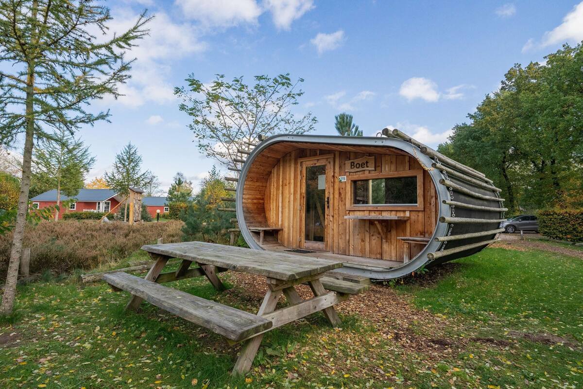 Une structure en bois en forme de bateau, dotée d’une porte portant l’inscription « Boet », d’un banc en bois et d’une table de pique-nique.