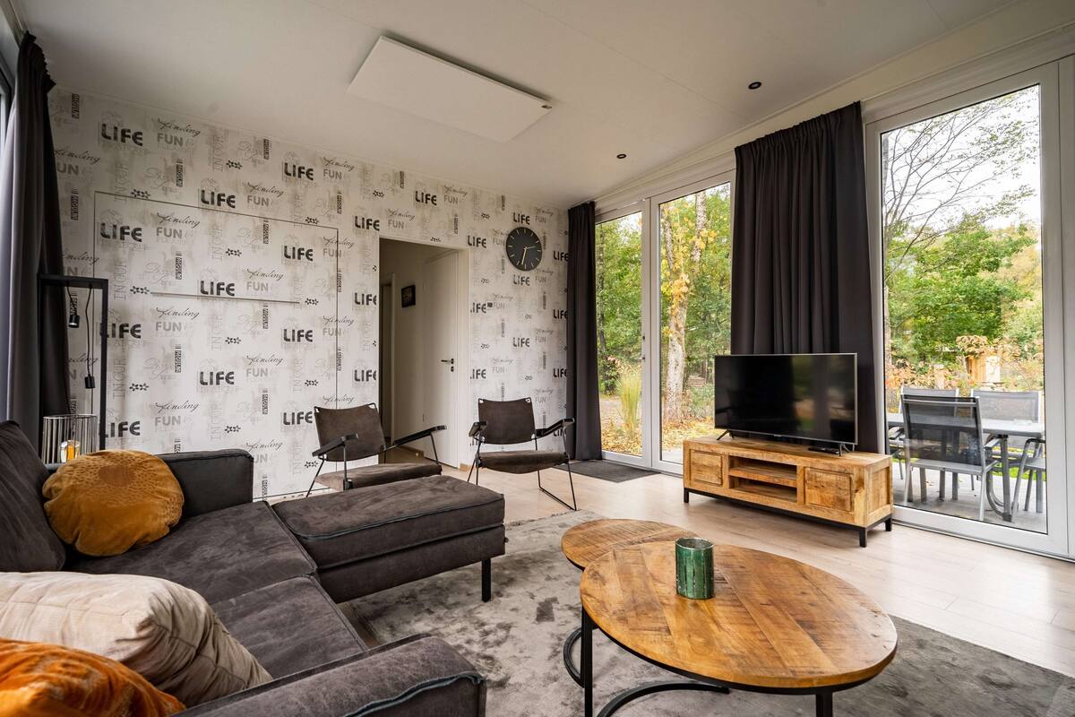 Un salon moderne comprenant un canapé, des fauteuils, une table basse, un téléviseur et une grande fenêtre donnant sur un paysage arboré.