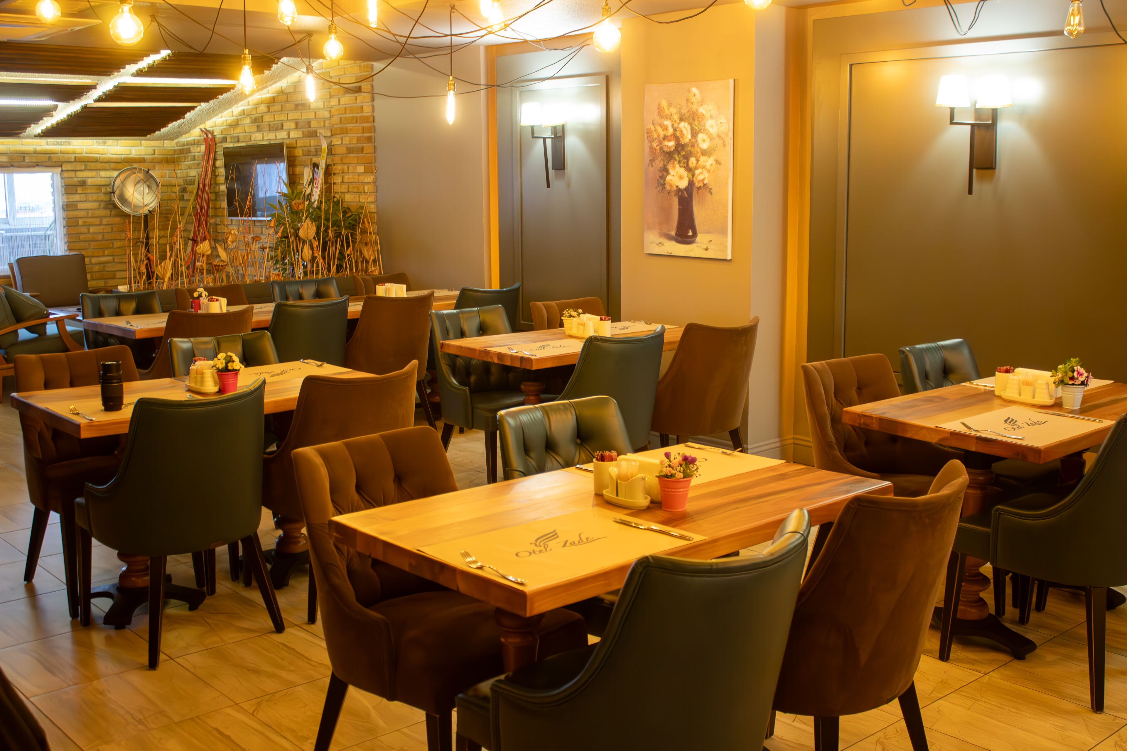 A restaurant with multiple wooden tables and chairs, a brick wall, and string lights.