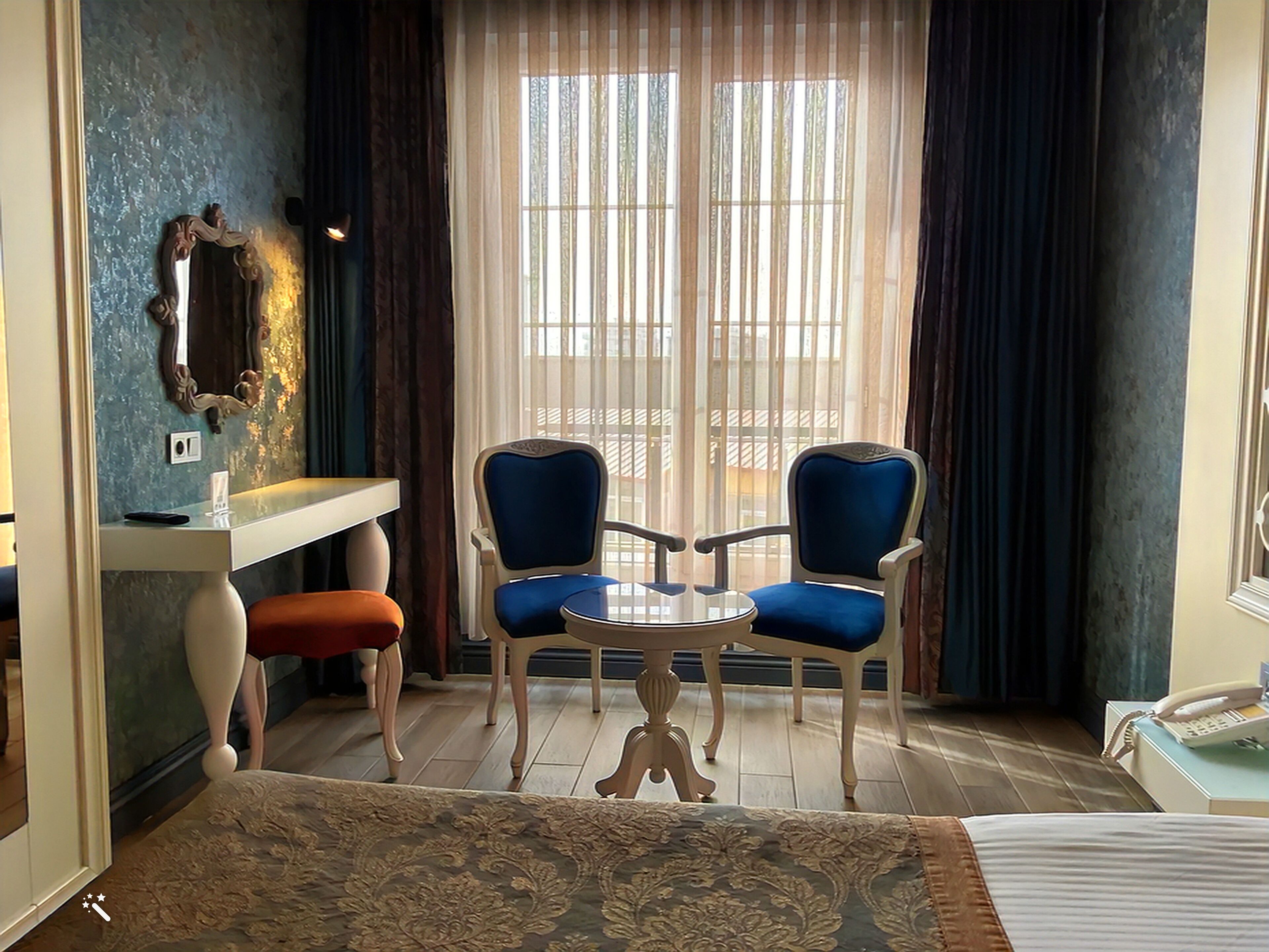 Habitación de hotel con una cama, un escritorio con espejo, dos sillas, una mesita y una ventana con cortinas.