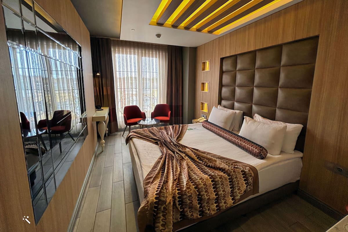 A modern hotel room with a large bed, a sitting area with two chairs, a small table, and a large window with curtains.