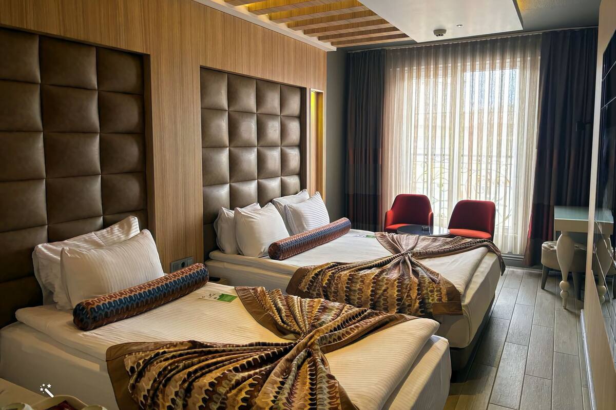 A hotel room with two beds, a large padded headboard, a view of the outdoors through sheer curtains, and a small table with a chair.