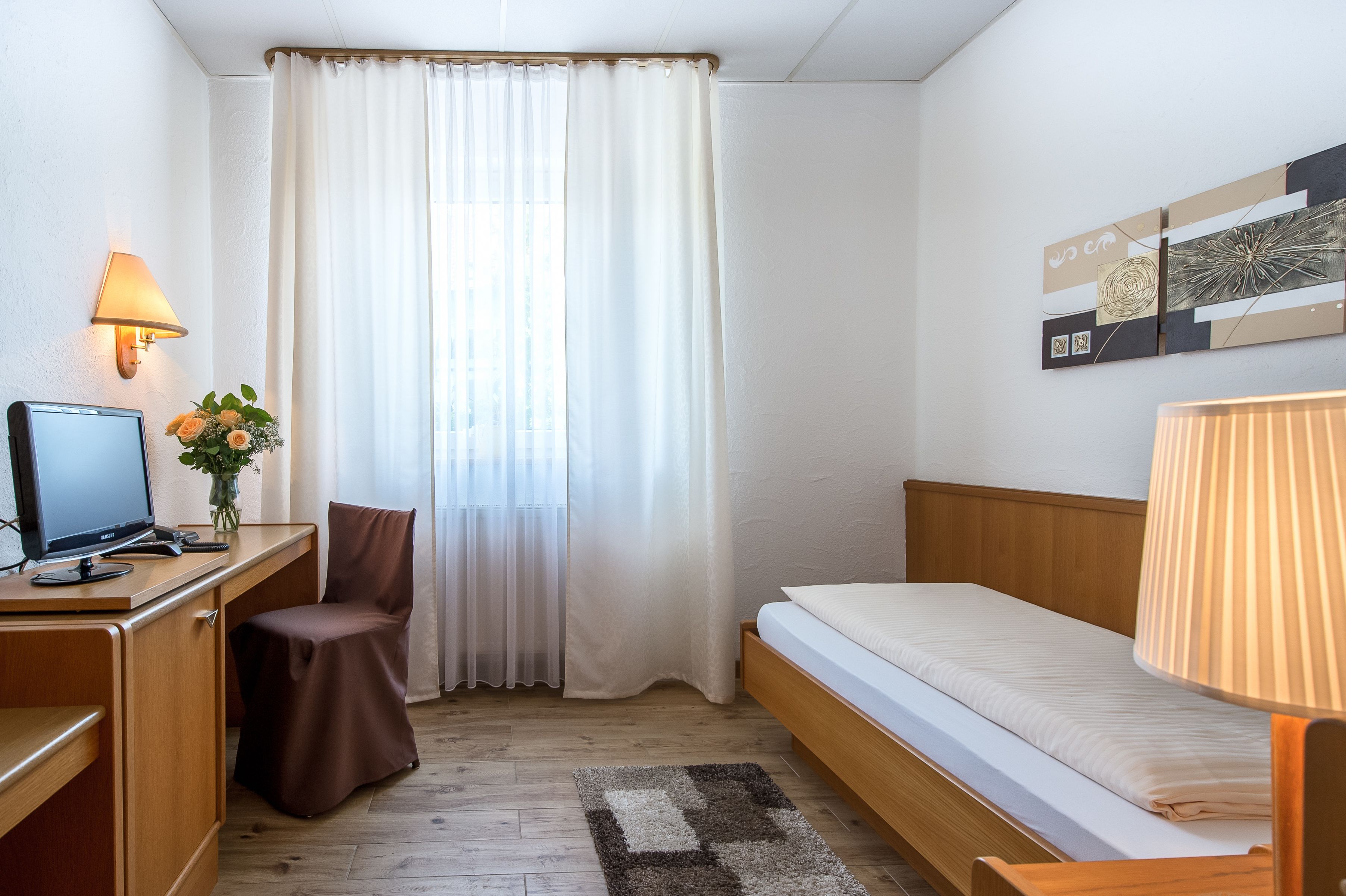 A hotel room with a wooden desk, a computer, a chair, a bed, and a window with white curtains.