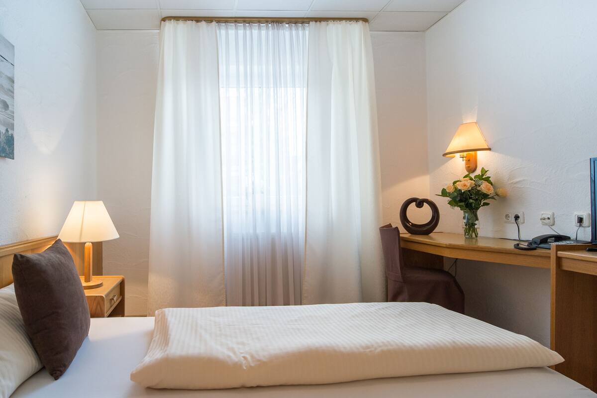 Ein Hotelzimmer mit Bett, Nachttisch mit Lampe, Schreibtisch ebenfalls mit Lampe, einer Vase mit Blumen und einem an der Wand befestigten Telefon.