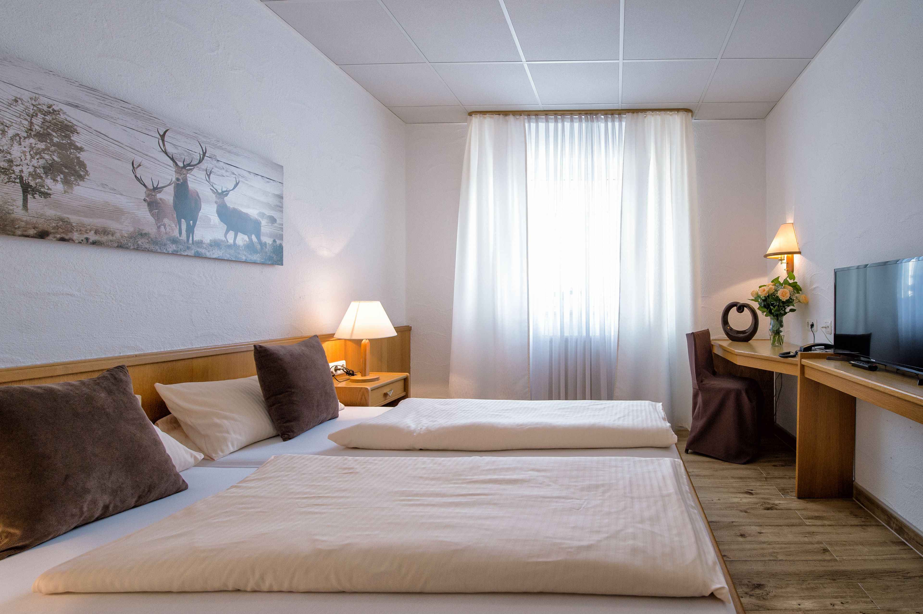 A hotel room with two beds, a desk with a TV, a lamp, and a painting of deer on the wall.