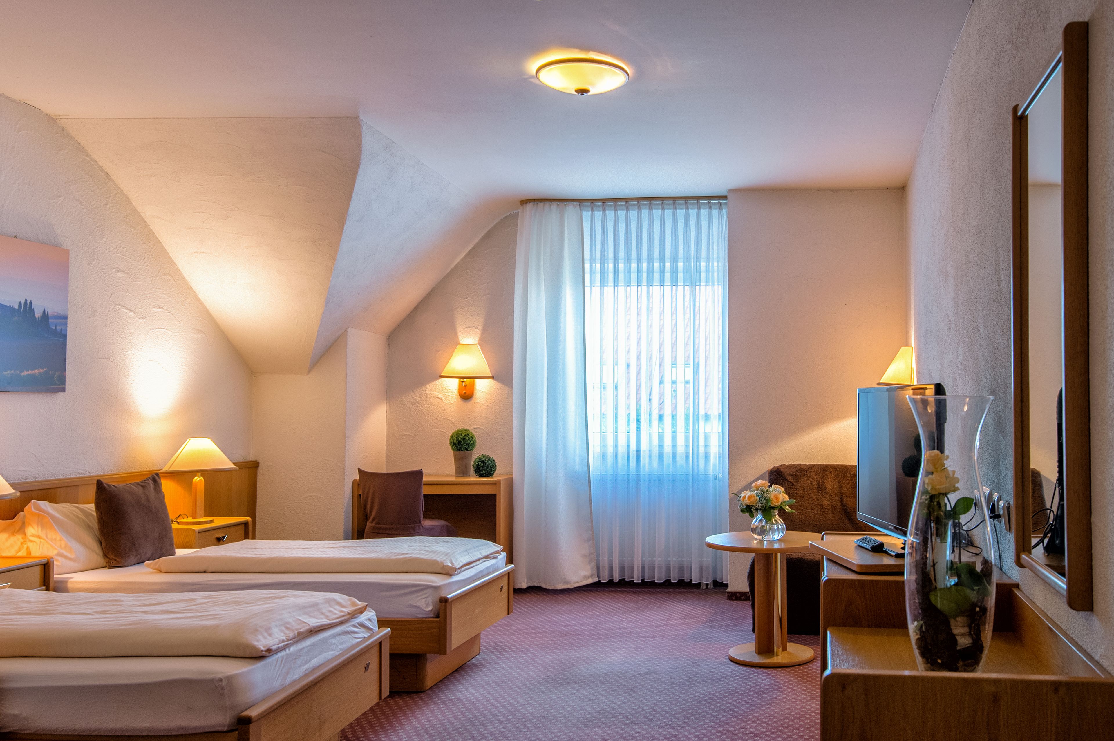 Una habitación de hotel con dos camas, un escritorio con computadora, un televisor y un florero con flores.