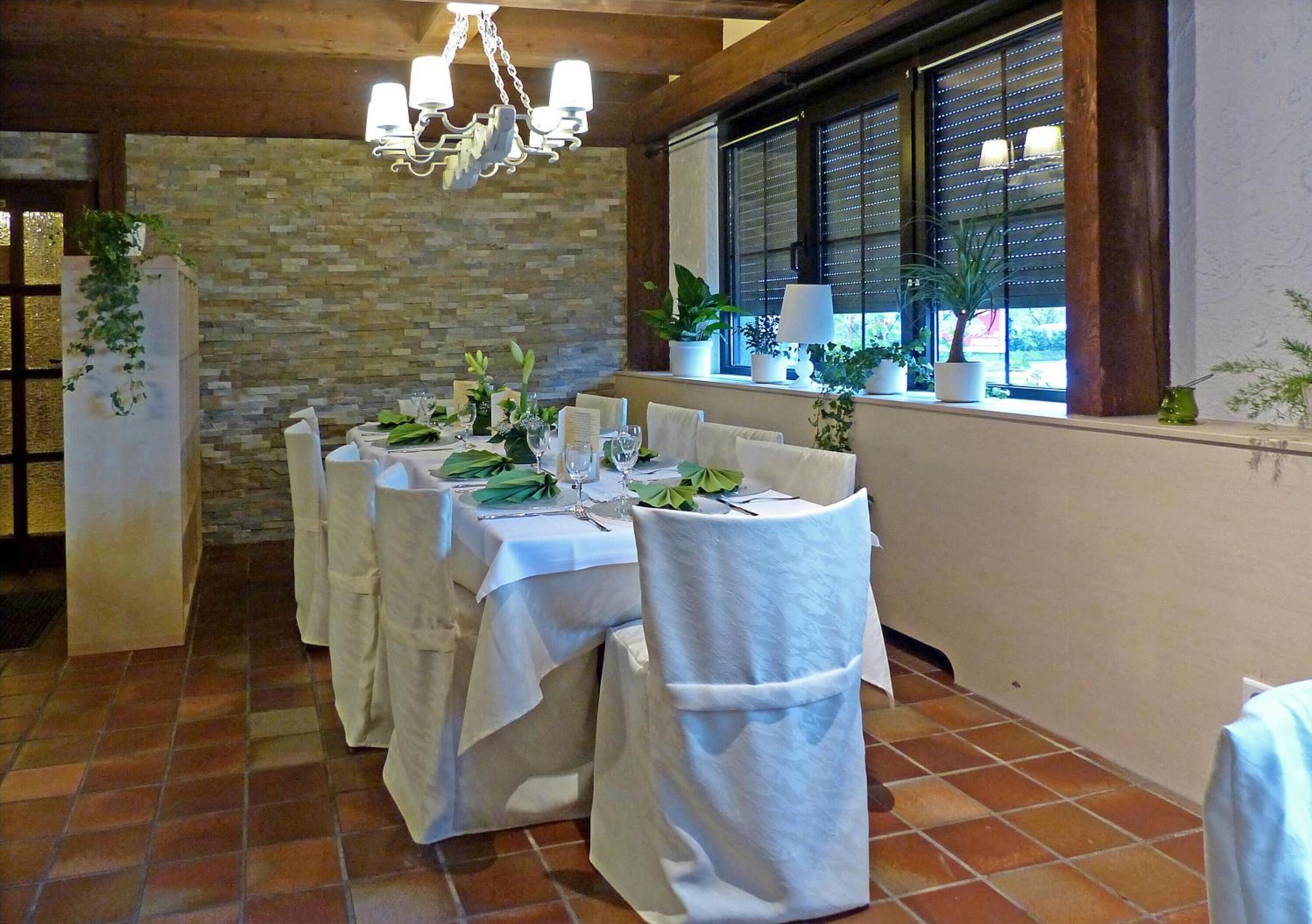 A dining area with a long table set for a meal, surrounded by chairs with white covers, a stone wall, and a window with blinds.