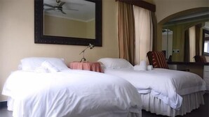 Habitación de hotel con dos camas, ventilador de techo, espejo y ventana con cortinas.