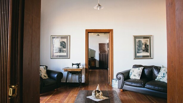 Un salón con piso de madera, una máquina de coser, un sofá de cuero negro y dos cuadros en las paredes.