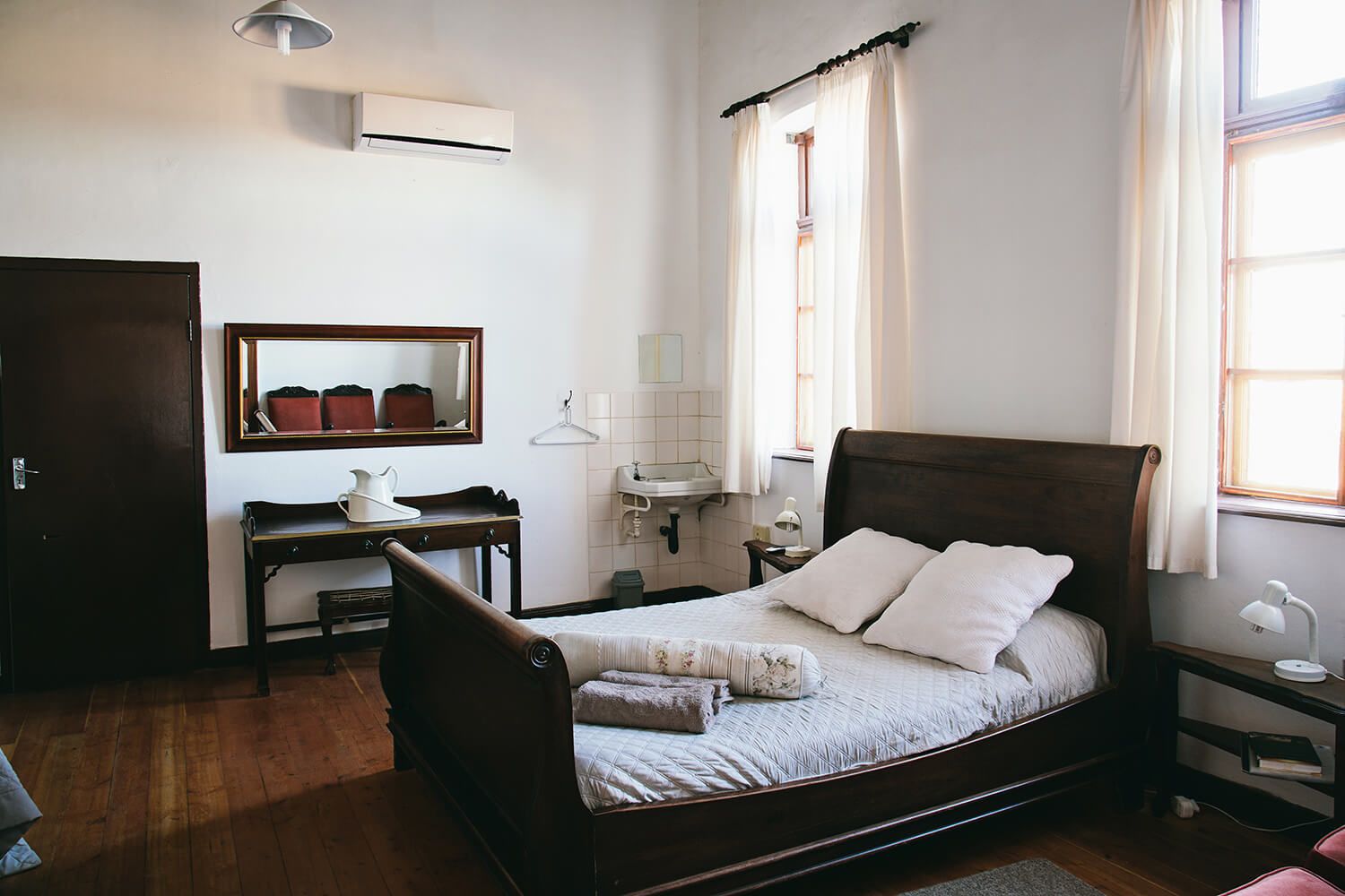 Een slaapkamer met een houten bed, een bureau met stoel, een spiegel, een raam met gordijnen, en een airconditioning.