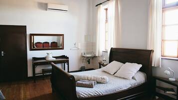 Een slaapkamer met een houten bed, een bureau met stoel, een spiegel, een raam met gordijnen, en een airconditioning.