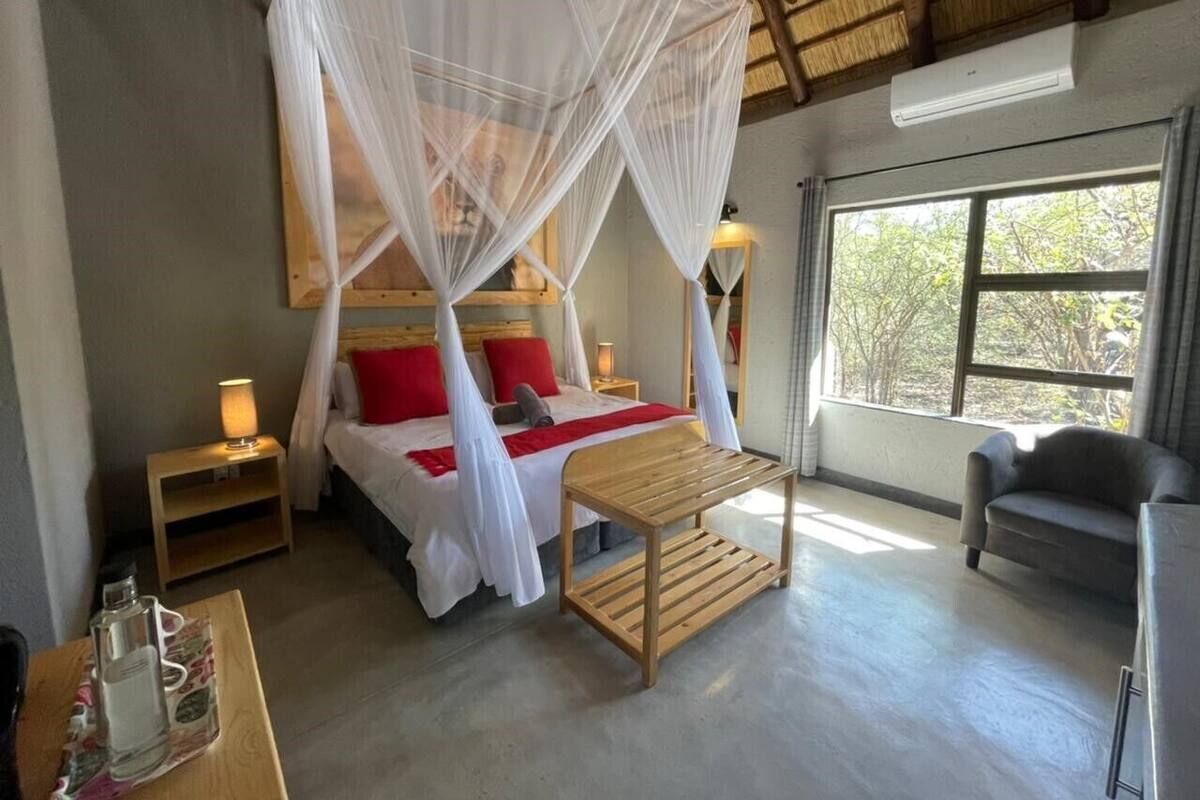 A bedroom with a bed under a canopy, bedside lamps, a wooden table, and a view of trees through a window.
