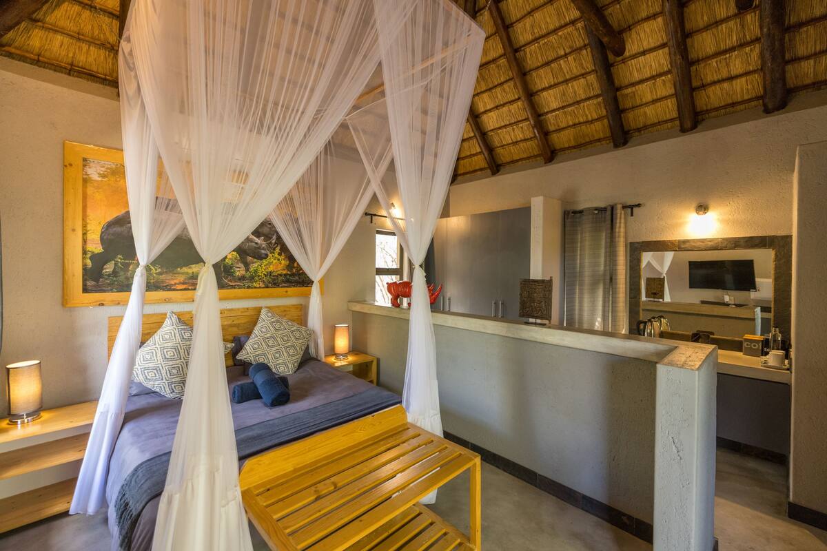 A bedroom with a wooden bed, a canopy, a painting, a bedside table with a lamp, and a view of a kitchen area.