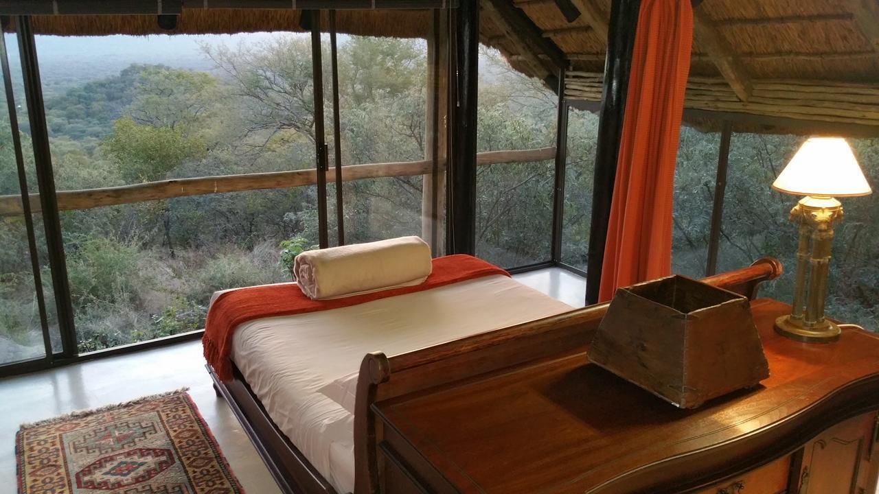 Une chambre avec une grande fenêtre, un lit recouvert d’une couverture rouge, une table de chevet en bois avec une lampe, et une vue sur l’extérieur.