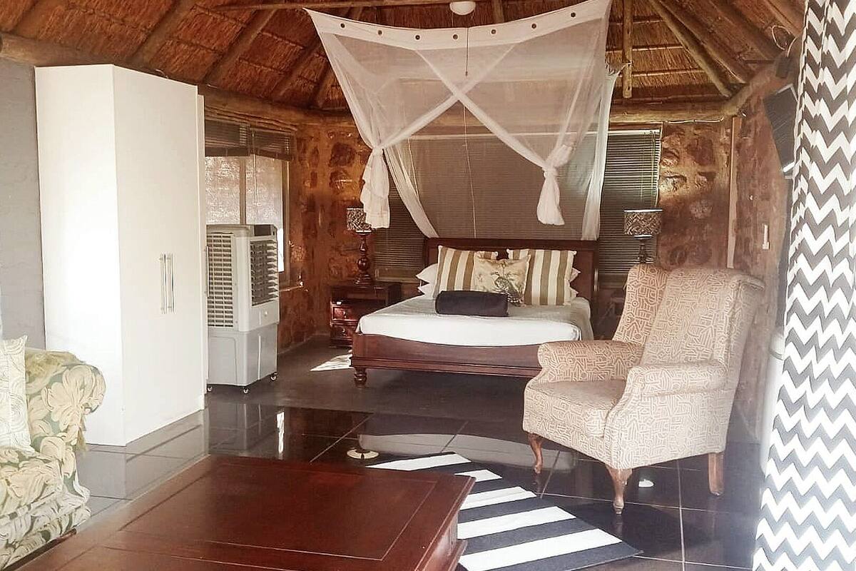 A bedroom with a bed, a chair, a bedside table, a wardrobe, a mosquito net, and a window with shutters.