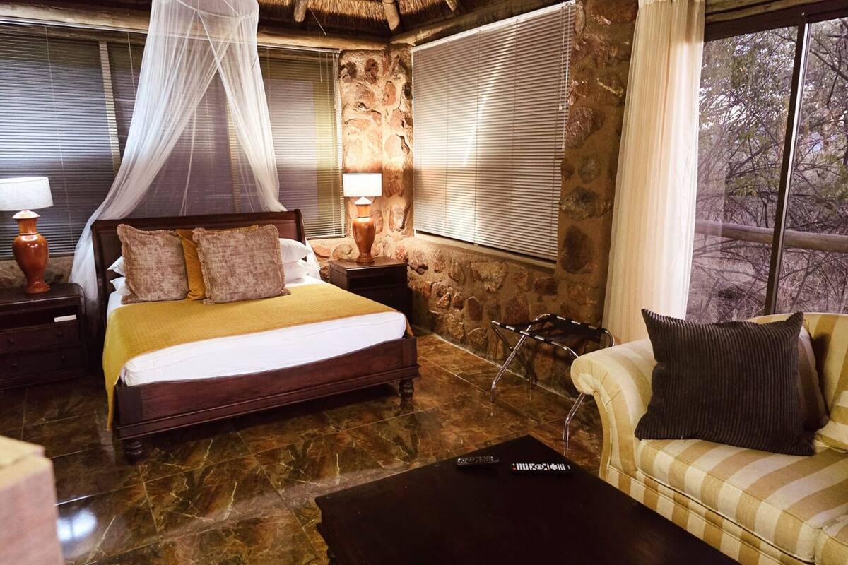 A bedroom with a bed, bedside lamps, a nightstand, a sofa, and a view of trees through the window.