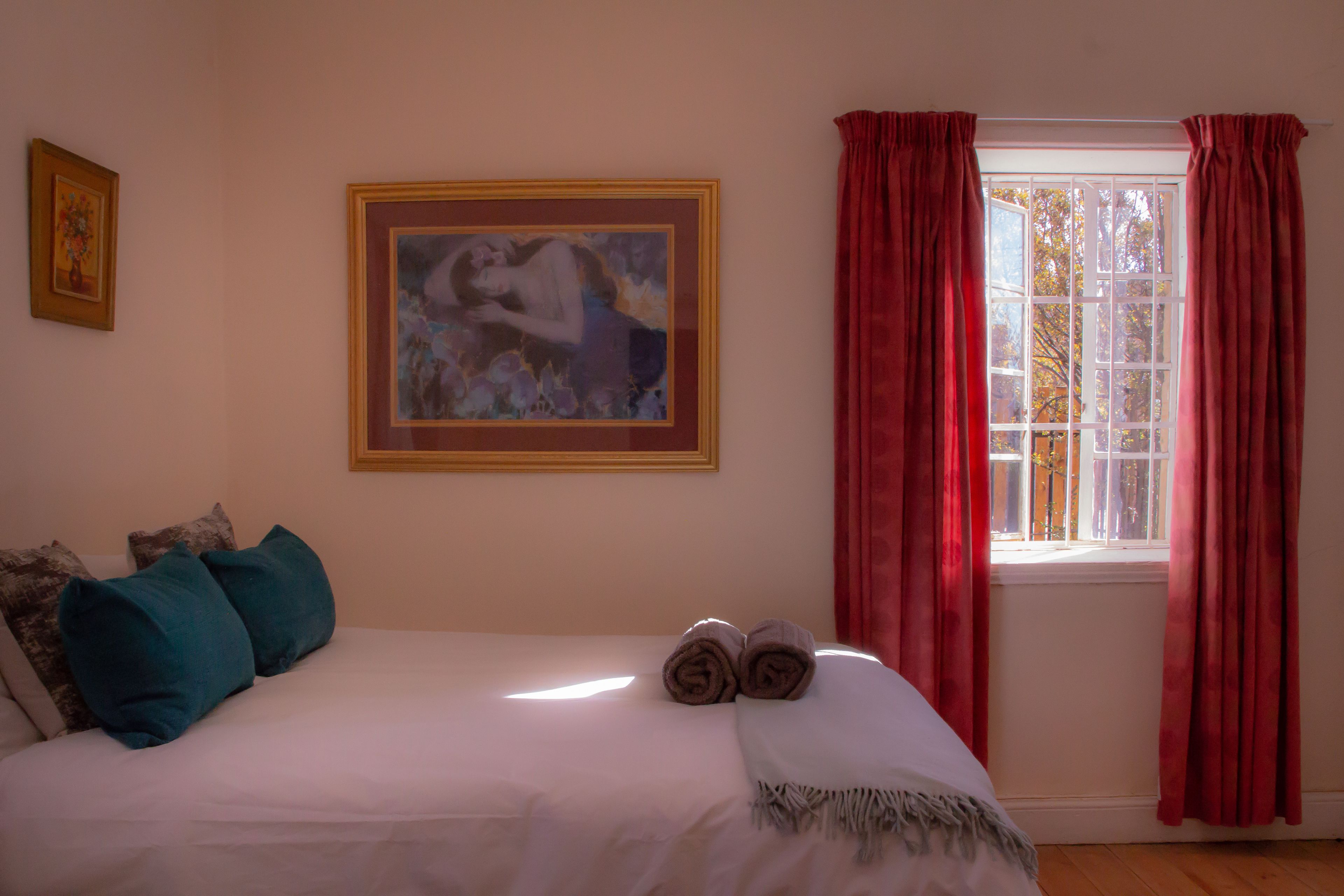 A neatly made bed with rolled towels, a framed painting on the wall, and a window with red curtains.