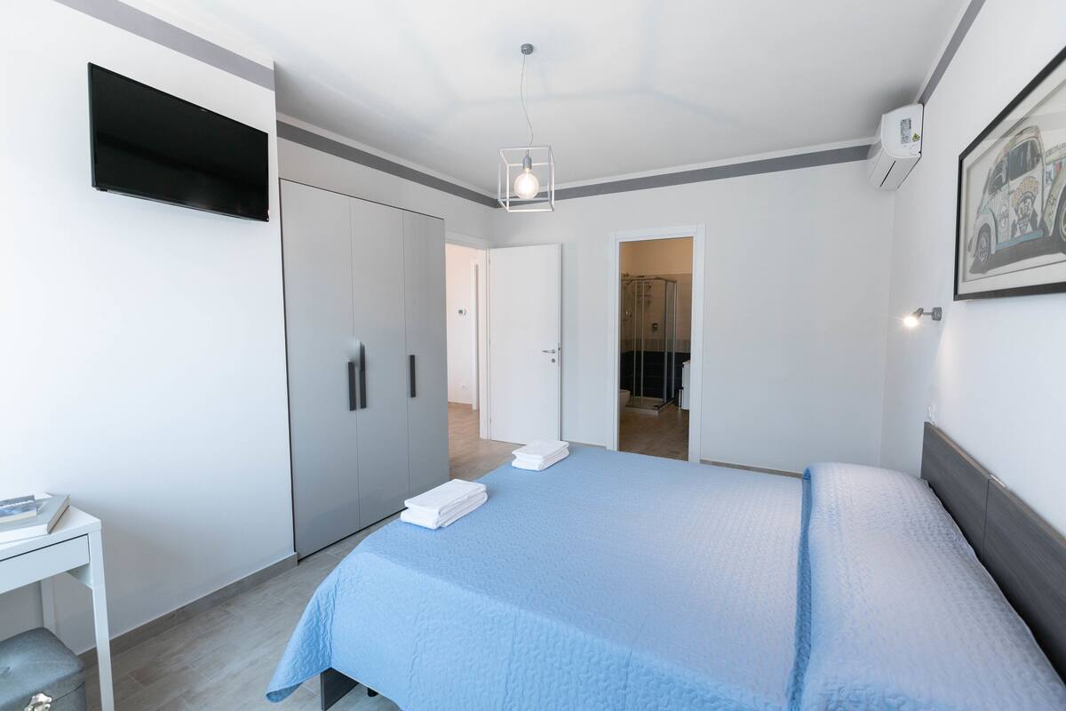 Un dormitorio moderno con cama, televisor montado en la pared, armario y baño visible a través de una puerta abierta.