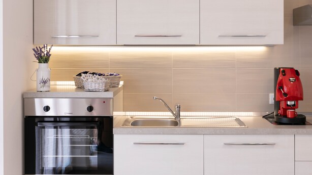 Una cocina moderna con armarios blancos, un horno de acero inoxidable, un lavamanos y una cafetera roja.