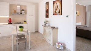 Una cocina moderna con armarios blancos, una mesa de comedor con sillas, una cafetera y una pequeña planta en maceta. Hay un cuadro en la pared y una cama en la habitación contigua.