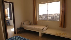 Una habitación pequeña con una cama individual, una ventana con cortinas, una silla y vista a un paisaje rocoso.