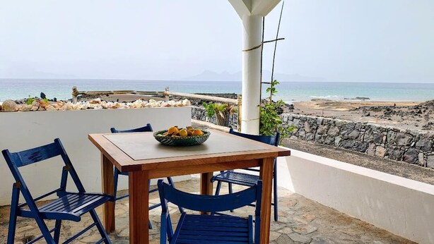 Una mesa de madera con sillas azules y un cuenco de fruta en un patio con vistas a la playa.