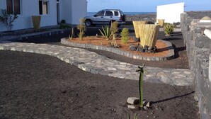 Un edificio blanco con un coche aparcado, un camino de piedra y un pequeño jardín con plantas.