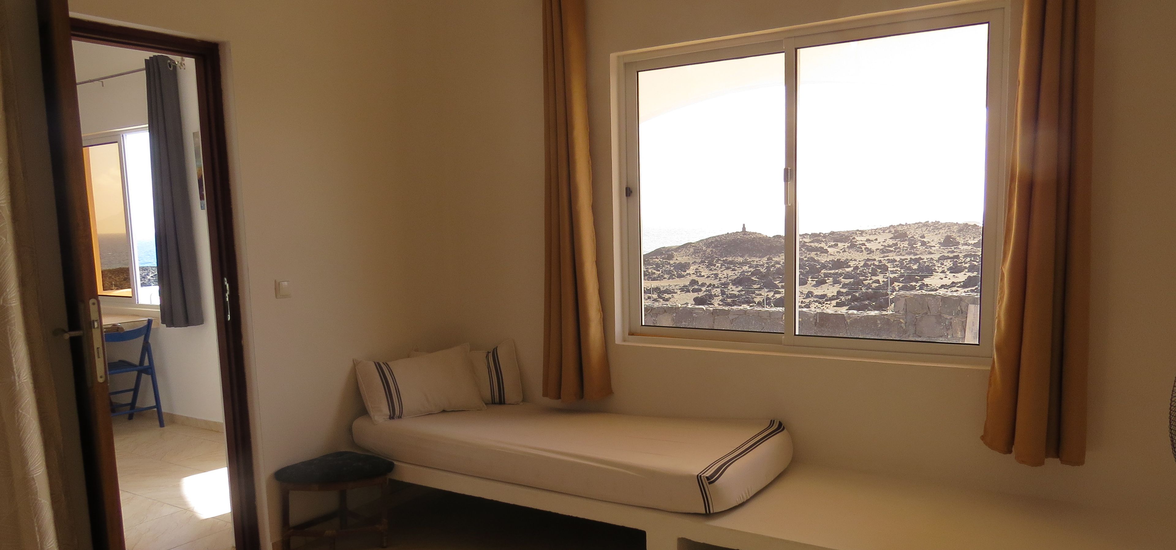 Une petite pièce avec un lit simple, une fenêtre avec des rideaux, une chaise et une vue sur un paysage rocheux.