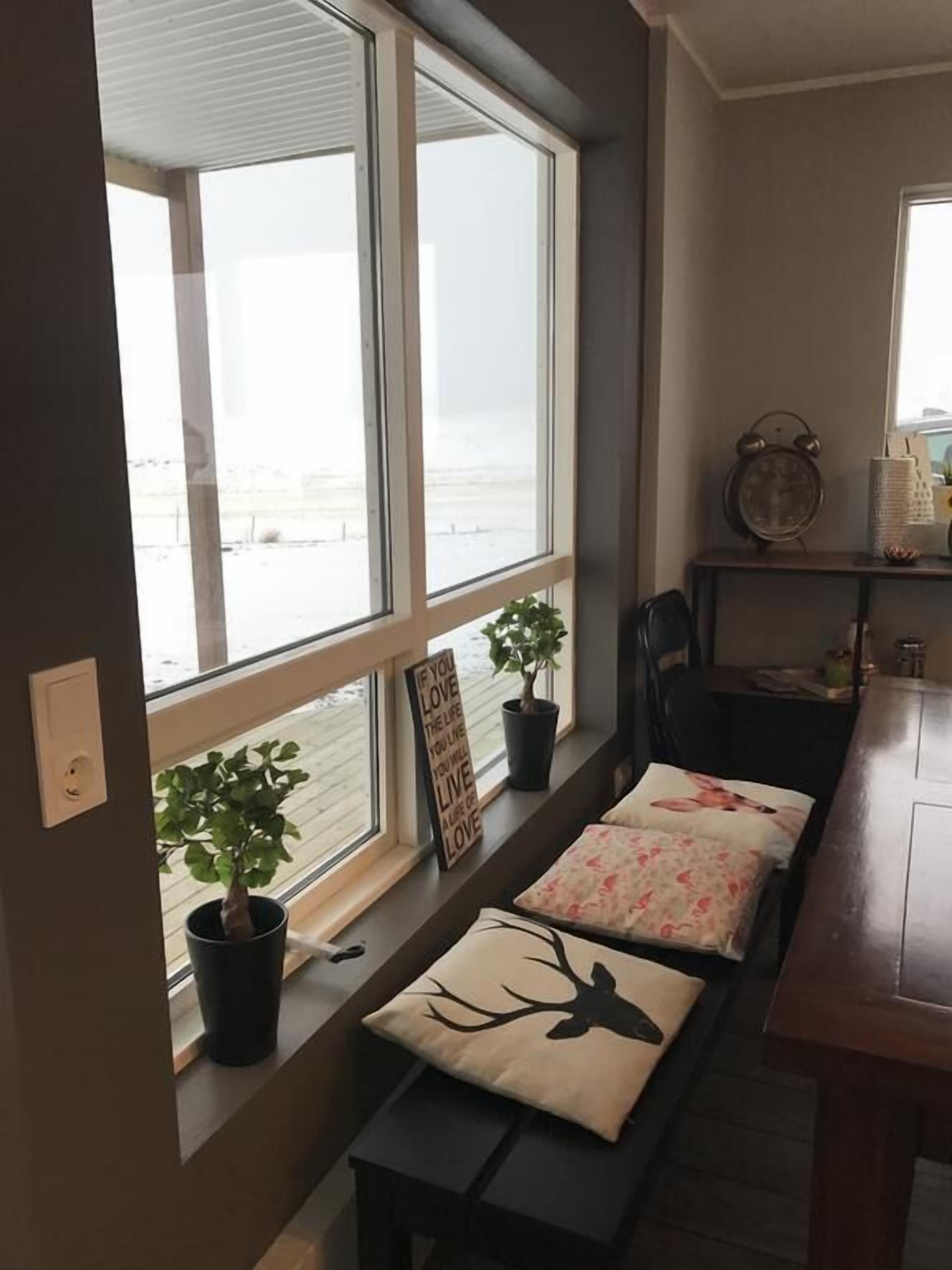 A room with a large window, a small table with a chair, a potted plant, and a decorative sign.