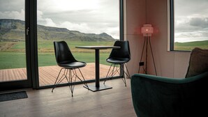 A room with a large window offering a view of a green landscape and mountains. There is a table with two chairs and a floor lamp. - Stóra Mörk Guesthouse (Rangárþing eystra)