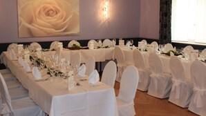 Un salón de banquetes con largas mesas dispuestas para un evento formal, sillas blancas y un gran cuadro floral en la pared.